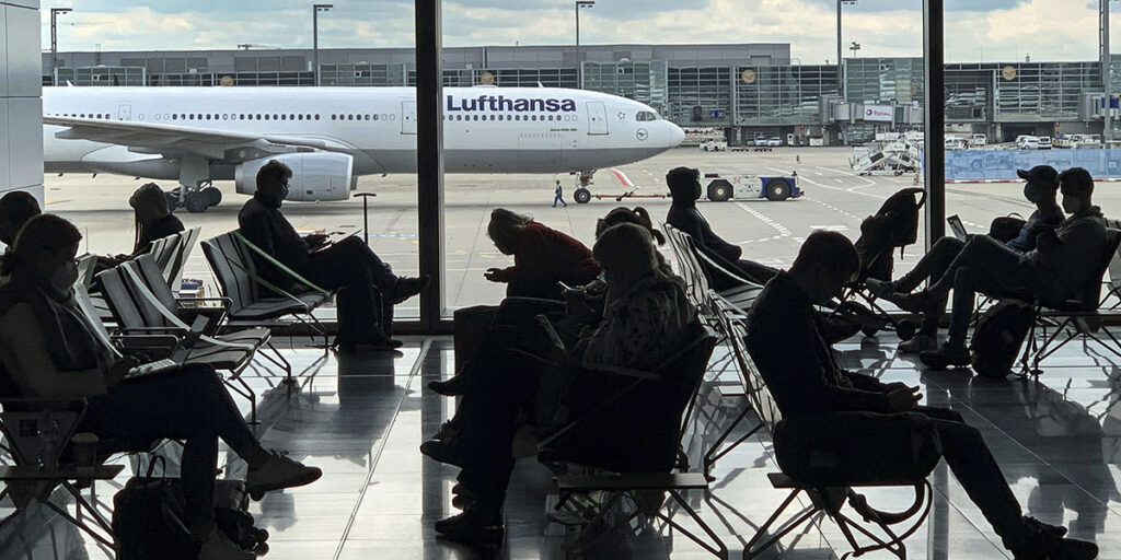Flygresenärer som sitter och väntar asvtecknas som siluetter mot ett stort fönster ut mot flygplatsen. I bakgrunden syns ett av flygbolaget Lufthansas vita passagerarplan.
